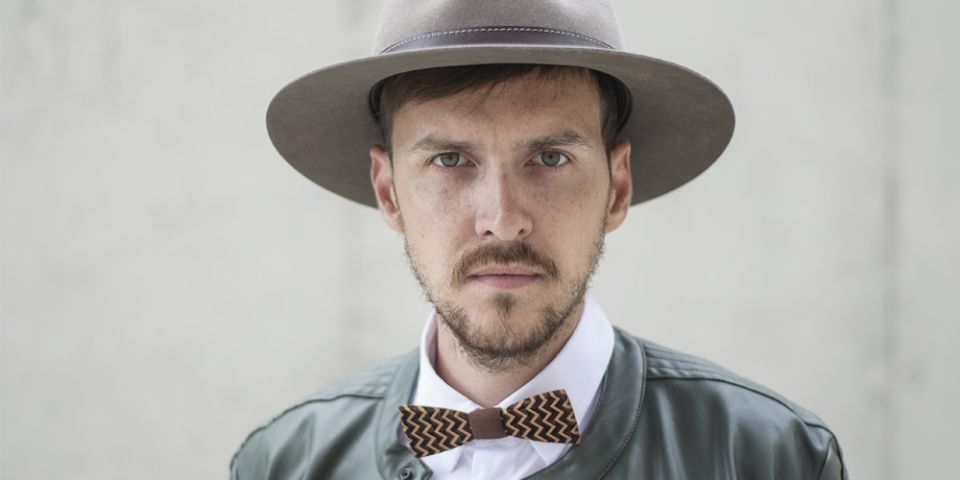 A man wearing brown hat with wooden detail, leather jacket and wooden bow tie. A man wearing brown hat with wooden detail, leather jacket and wooden bow tie.